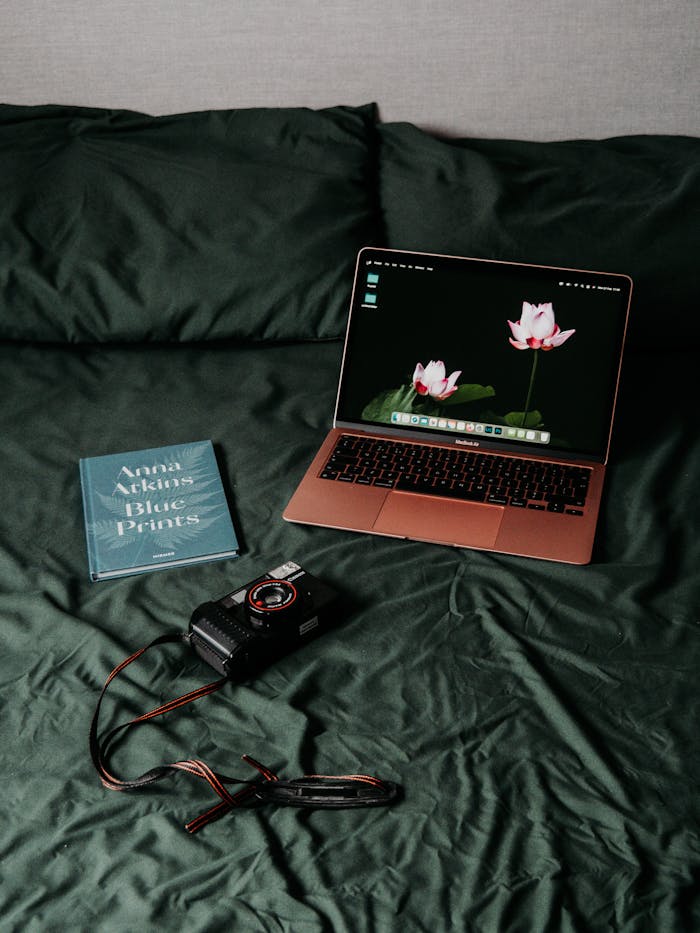 A cozy home workspace featuring a laptop, camera, and book on a bed. Ideal for creative professionals.